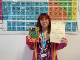 Student Bea stands in front of a Periodic Table poster. She is holding her certificate and prize