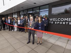 CEO of Loughborough College Group Corrie Harris and Mayor of Charnwood David Northgate cut the ribbon in front of the new building. There are people behind them to the left and a sign saying WELCOME TO SPORTS on the wall behind them on the right