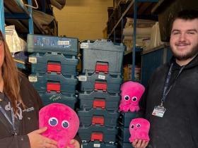 Staff hold pink fluffy octopus toys and stand in front of a stack of tools