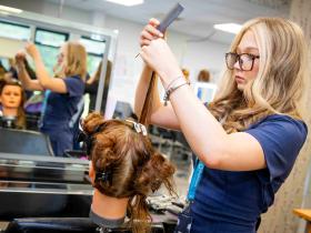 A hairdressing student works on cutting hair on a mannequin in a salon. She is stood in front of a mirror.