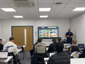 Ben stands at the front of a classroom. Students are sat at desks with their laptops. The screen has a presentation of rugby analysis
