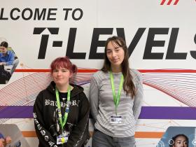 Jess and Ella stand in front of a wall with pictures of students working. The wall reads: Welcome to T Levels