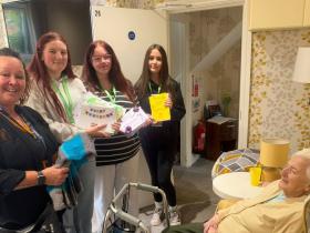 Curriculum Head Sharon with students holding cards and gifts for Cissy, sat in a chair on the right hand side
