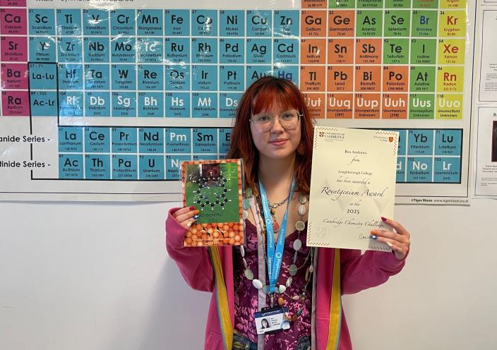 Student Bea stands in front of a Periodic Table poster. She is holding her certificate and prize