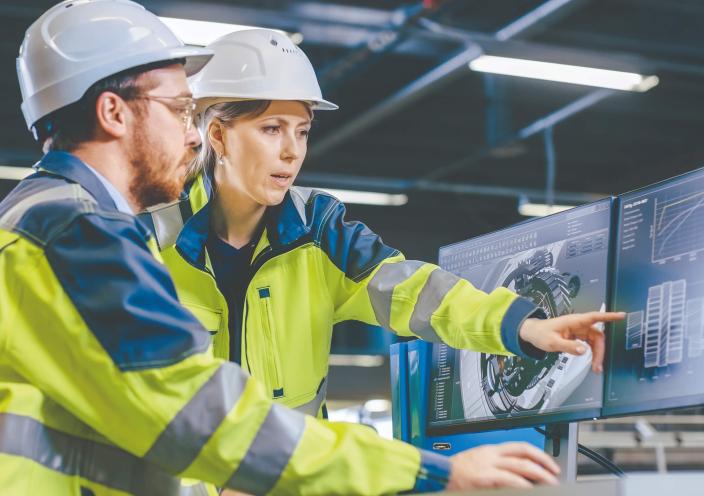 A female engineer pointing at a computer screen while talking to a male engineer who is working on the computer, both are wearing high vis jackets and hard hats.