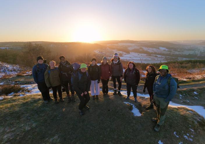 A group of walkers on a hill in the Peak District. There is snow around them and the sun is rising behind them