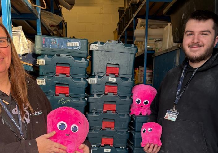 Staff hold pink fluffy octopus toys and stand in front of a stack of tools