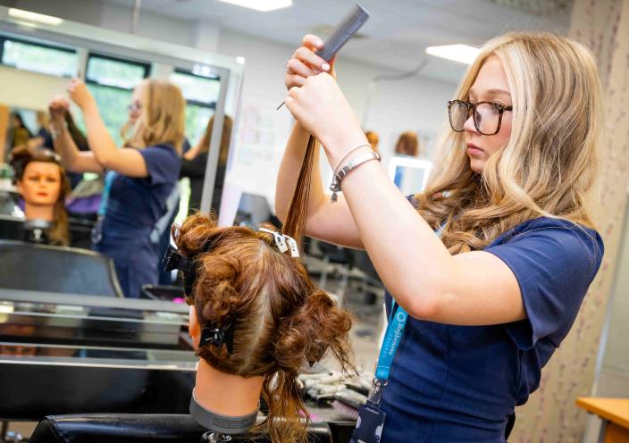 A hairdressing student works on cutting hair on a mannequin in a salon. She is stood in front of a mirror.