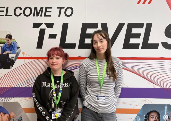 Jess and Ella stand in front of a wall with pictures of students working. The wall reads: Welcome to T Levels