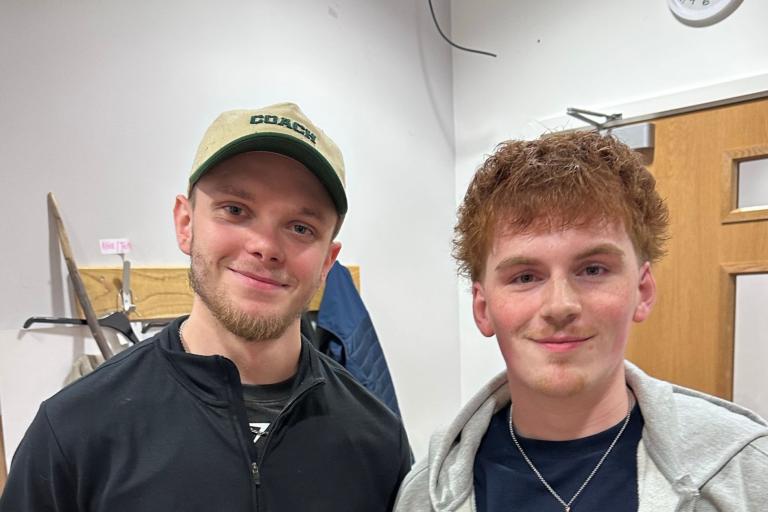 Colby and Dom stand together smiling at the camera. One is wearing a hat.