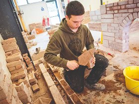 A construction student holding a brick and adding cement to it with a trowel before adding to the wall they're building