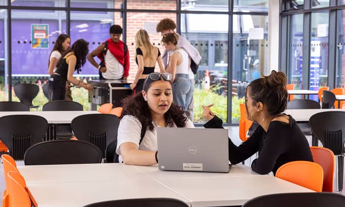 Student in the 1909 Lounge, in the background a group are playing table football while in the foreground two students are sitting at a table chatting while looking at a laptop computer