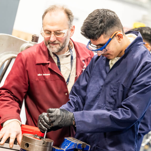 Apprentice in overalls and safety goggles standing in a workshop being guided by a tutor