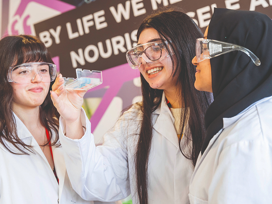Group of students wearing lab coats and safety goggles, holding up a petri dish