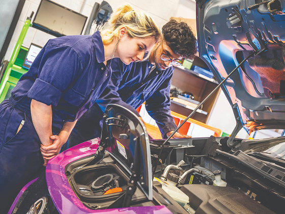 Two people in blue overalls, looking into the open bonnet of a car