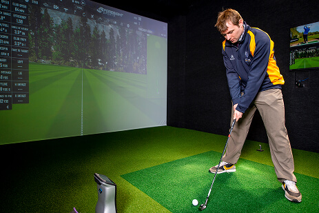 A man standing in the gold simulator with a gold club in a stance that shows he's ready to swing and hit the gold ball