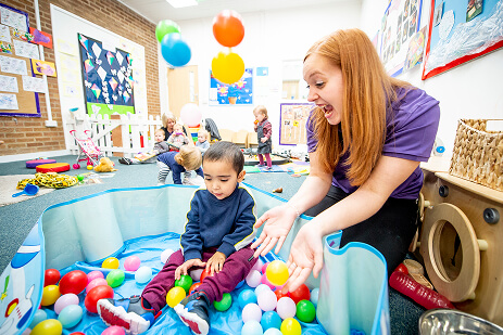 Child sitting in a ball pit throwing balls in the air. A nursery staff member is beside the ball pit. Other children are playing in the background.
