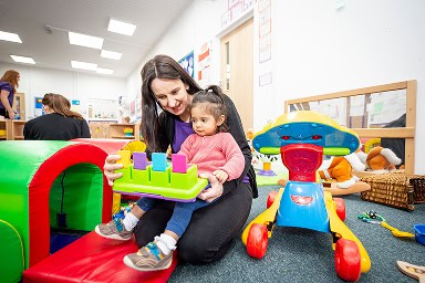 Inside a day care nursery a care worker is sitting with a child on her knee and playing with a bright coloured toy