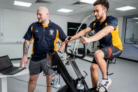 An athelete on an exercise bike indoors in a lab with a student monitoring their vital signs live on a computer