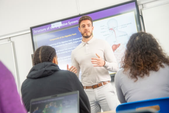 Undergraduate lecturer standing in front of a class teaching students with a white board behind them