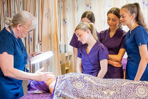 A group of beauty therapy students standing watching their tutor perform a face massage on a volunteer customer